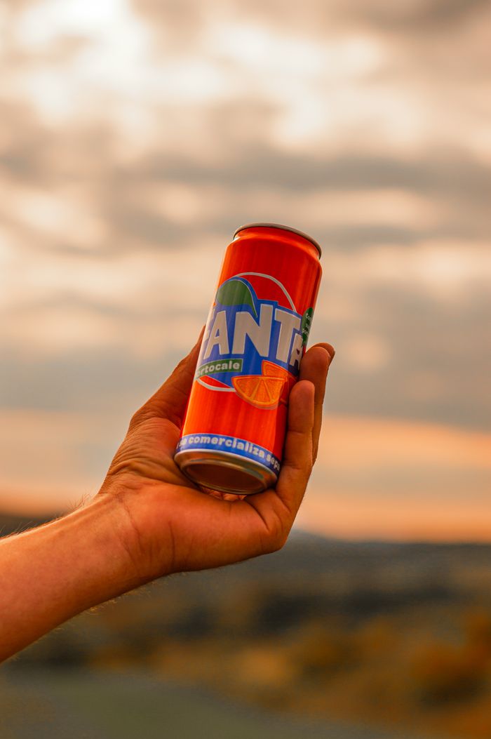 services-02 A hand holds a can of orange soda against a blurred outdoor background with a warm atmosphere.